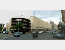 Ampio incrocio urbano con una lunga struttura beige a più piani e, sul lato sinistro, un parcheggio multipiano circolare con rampe esterne e ringhiere rosse. Lungo la strada si vedono auto, semafori e palazzi residenziali sullo sfondo.