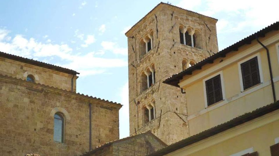 Torre campanaria in pietra chiara con finestre ad arco e colonnine, che svetta tra edifici storici con tetti in coppi e facciate intonacate. Sullo sfondo cielo azzurro con leggere nuvole.