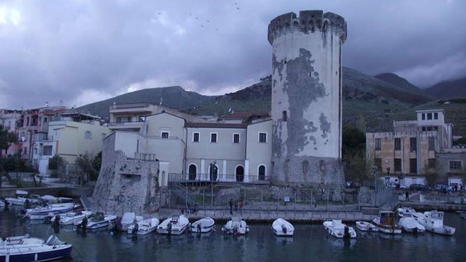 Porticciolo con piccole barche ormeggiate lungo la banchina e una torre costiera cilindrica in pietra con sommità merlata. Sullo sfondo si vedono edifici bassi del paese e colline montuose sotto un cielo nuvoloso.