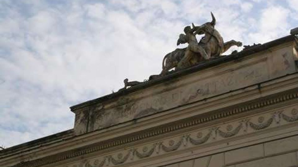 Facciata superiore di un edificio in stile classico con cornici e fregi decorativi in pietra, ripresa dal basso. Sul tetto è visibile un gruppo scultoreo con cavallo e figure, stagliato contro un cielo parzialmente nuvoloso.