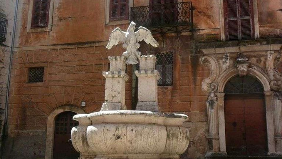 Fontana in pietra al centro di una piccola piazza, con vasca circolare su gradoni e scultura di un’aquila su piedistalli. Sullo sfondo si vede la facciata di un edificio storico intonacato, con persiane scure, balconcino in ferro e portale ad arco decorato.