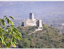 Castello in pietra con torre centrale e mura merlate su una collina boscosa, con sentiero d’accesso visibile. Sullo sfondo si estende una valle con campi e insediamenti urbani leggermente velati dalla foschia.