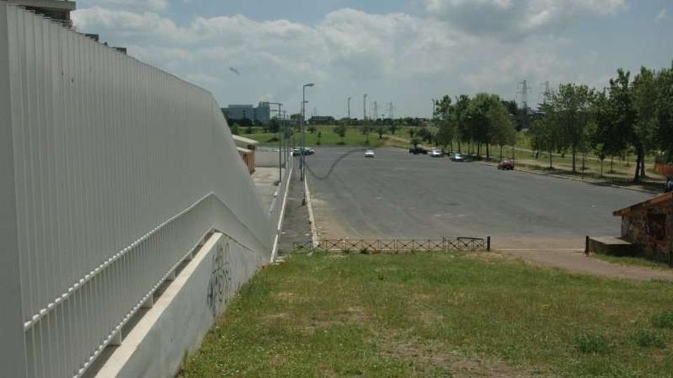 Ampio parcheggio asfaltato quasi vuoto accanto a una lunga struttura moderna con facciata in pannelli metallici e corrimano. Intorno ci sono prati, una strada con lampioni e filari di alberi, con cielo parzialmente nuvoloso sullo sfondo.