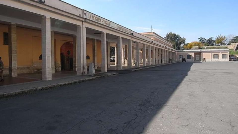 Cortile asfaltato di un complesso a un piano con un lungo porticato laterale sostenuto da colonne rettangolari e pareti color ocra. Sul cornicione corre una scritta e, sullo sfondo, si vedono altri edifici bassi con alcune alberature.
