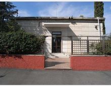Facciata di un piccolo edificio moderno a un piano, rivestito in pietra chiara, con ingresso centrale a porta vetrata sotto una pensilina. Davanti c’è un camminamento con muretto basso rosso e una grata in legno, circondato da siepi e alberi.
