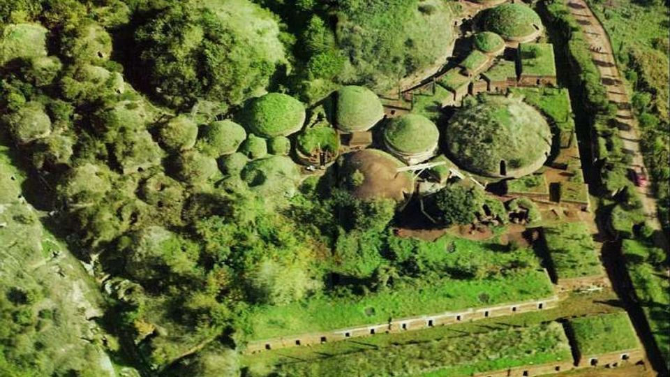 Vista aerea di un complesso immerso nel verde con numerose strutture circolari a cupola ricoperte d’erba, collegate da sentieri e muretti. Intorno si vedono alberi, aree terrazzate e un lungo muro di recinzione che delimita la zona.