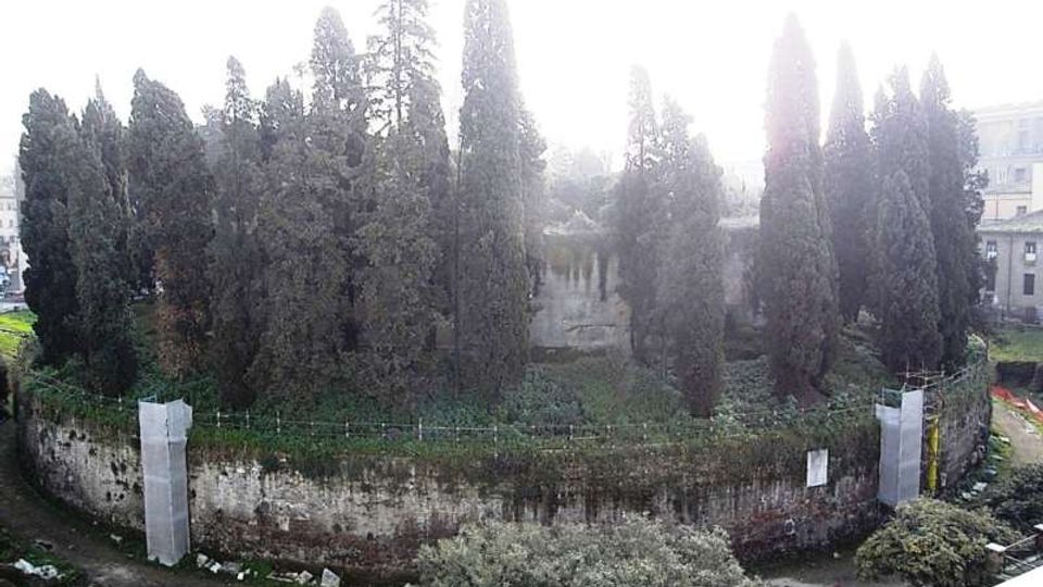 Area verde urbana con alti cipressi e altra vegetazione, circondata da un muro in pietra curvo con tratti di recinzione. Sullo sfondo si intravedono edifici residenziali e un piccolo corso d’acqua o fossato ai piedi del muro.