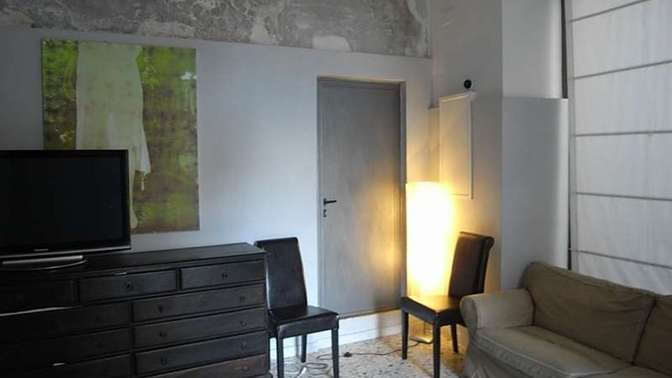 Interno di una stanza in stile moderno-industriale con pareti chiare e una fascia di intonaco grezzo vicino al soffitto. Sono visibili una cassettiera scura con TV e quadro, una porta centrale, due sedie nere, una lampada da terra luminosa e un divano accanto a una grande finestra con tenda.