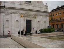 Facciata in pietra chiara di una chiesa monumentale con grande portale centrale, cornici scolpite e iscrizione lungo il fregio. In primo piano una piazza pavimentata con muretti bassi e una vasca d’acqua rettangolare; sullo sfondo edifici urbani e lampioni.