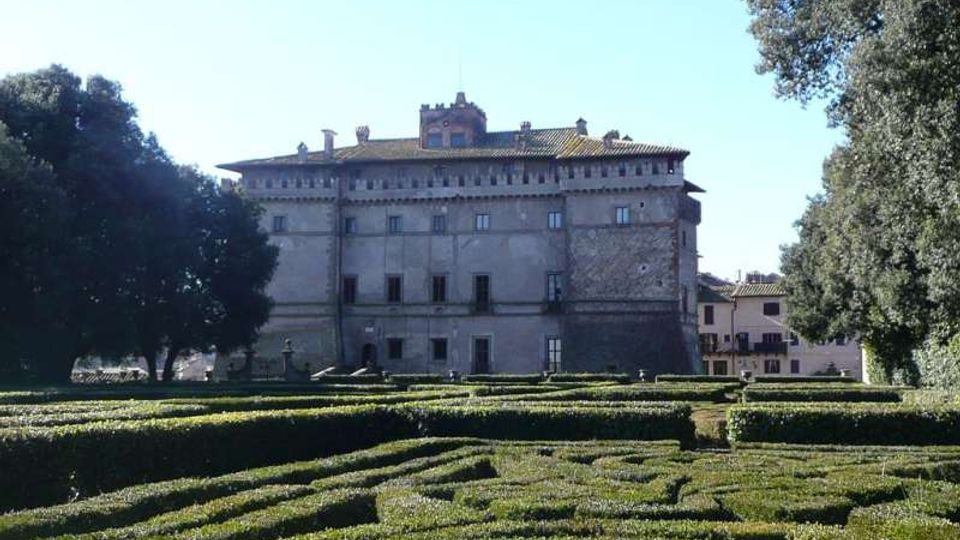 Veduta di un grande palazzo storico in pietra con tetto in coppi e finestre regolari, sullo sfondo di un cielo sereno. In primo piano si estende un giardino formale con siepi basse potate a labirinto, circondato da alberi ad alto fusto.