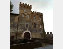 Facciata in pietra di un edificio storico fortificato con merlature, arco d’ingresso e finestre ad arco, con un piccolo orologio sulla parte alta. In primo piano si vede un muro di cinta e un vialetto in pietra sotto un cielo nuvoloso.