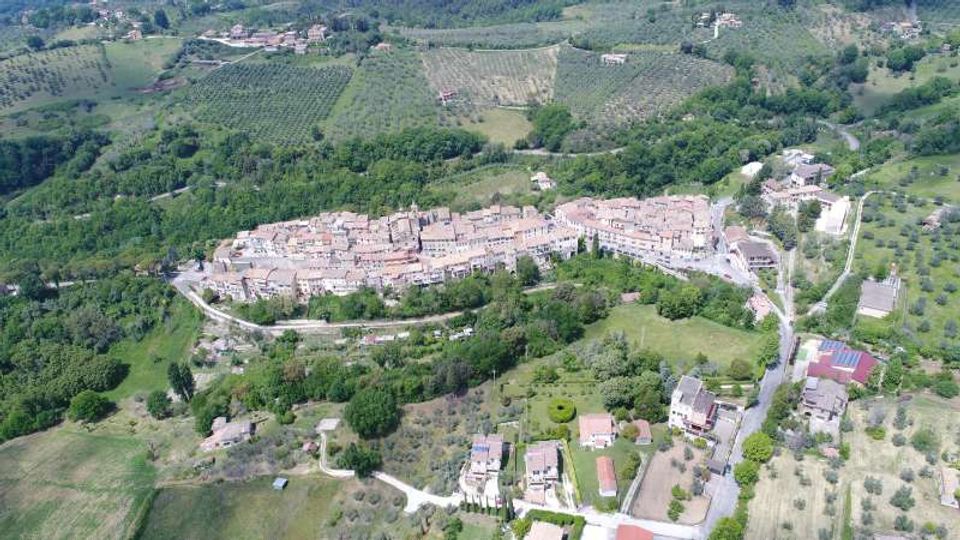 Veduta aerea di un borgo collinare con case in pietra addossate e tetti in coppi, circondato da boschi, campi coltivati e uliveti. Strade tortuose e abitazioni sparse si diramano dal centro abitato nella campagna circostante.
