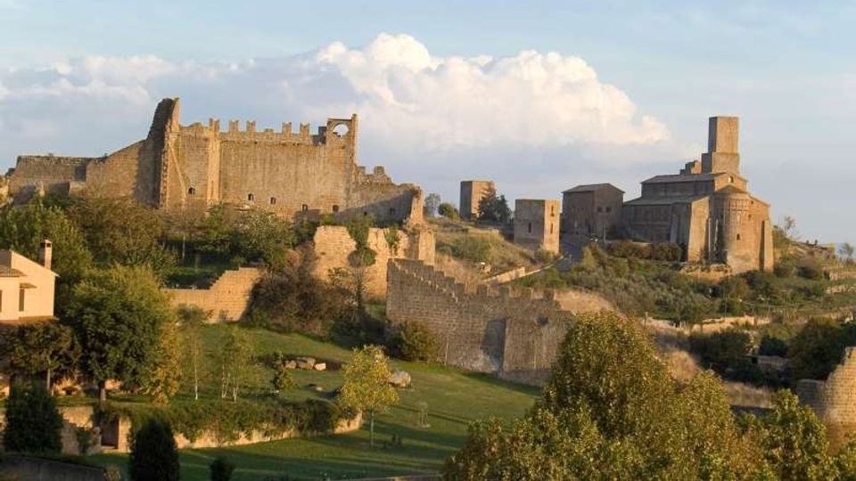 Panorama collinare con un castello medievale in pietra e mura merlate sulla sinistra e una chiesa in pietra con torre campanaria sulla destra. In primo piano si vedono bastioni e mura di cinta tra aree verdi, alberi e sentieri.