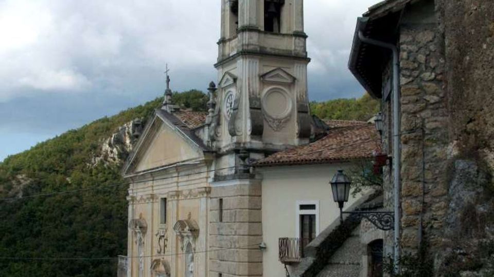 Chiesa storica in pietra e intonaco con facciata decorata e campanile con cupola scura, affacciata su una piccola piazza con fontana. Accanto corre una stretta strada lastricata lungo un muro in pietra, con colline boscose sullo sfondo sotto un cielo nuvoloso.