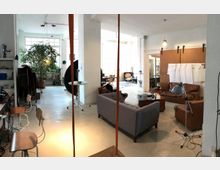 Ampio loft open space con grandi finestre e pilastri bianchi, con area salotto composta da divani in pelle e in tessuto, tavolino e lampade. Sullo sfondo si vedono postazioni di lavoro, scaffali e una grande pianta in vaso, con una porta scorrevole su binario.