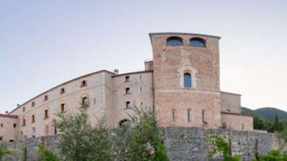 Grande complesso in pietra tipo castello o monastero su un basamento murario, con torre cilindrica e finestre ad arco, su una collina con vegetazione e montagne sullo sfondo sotto un cielo sereno.