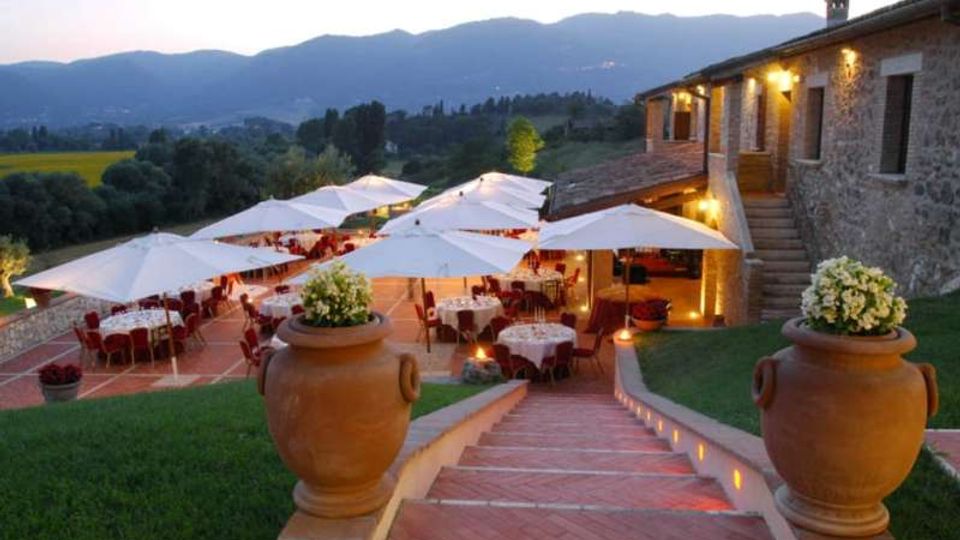 Terrazza all’aperto con tavoli e sedie sotto grandi ombrelloni bianchi, accanto a un edificio in pietra illuminato e raggiungibile tramite una scalinata in cotto. Sullo sfondo si vedono colline e montagne, con grandi vasi decorativi ai lati delle scale e aree verdi intorno.