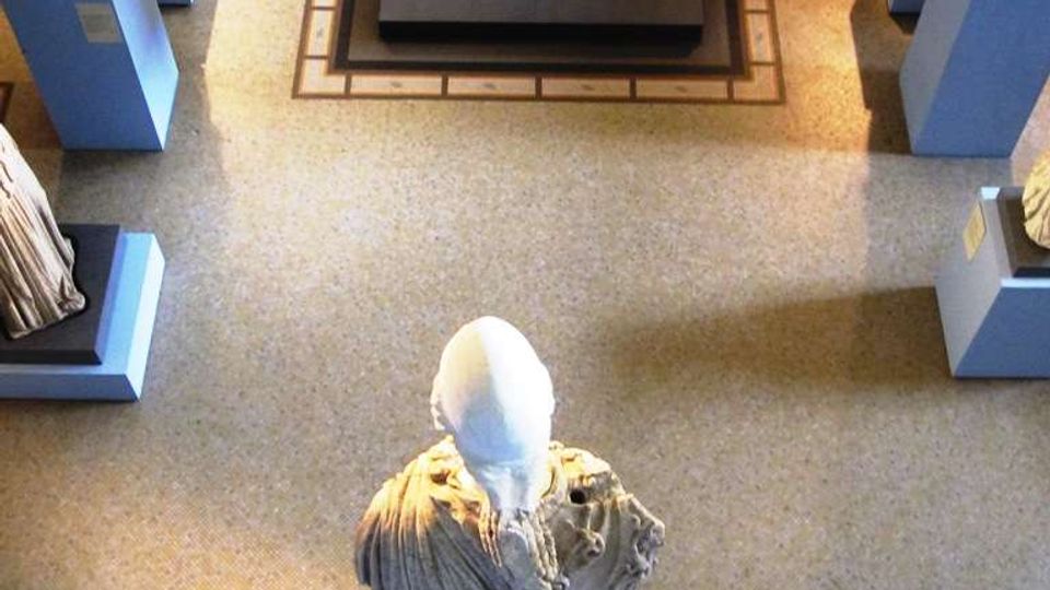 Interno di un museo visto dall’alto, con statue su piedistalli lungo i lati e una scultura centrale in primo piano su base quadrata. Sul fondo si notano una ringhiera in metallo e una scala che conduce a un livello superiore.