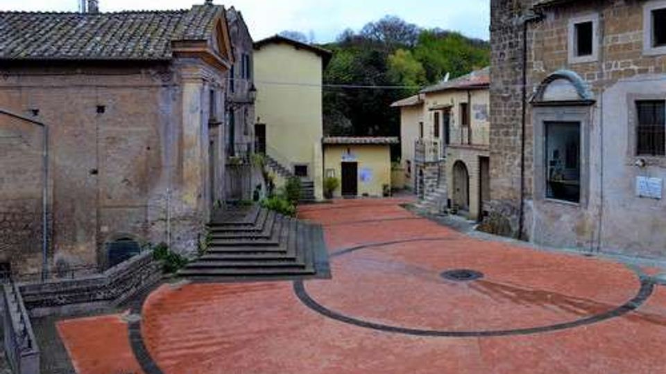 Piccola piazza lastricata in mattoni rossi con linee curve scure, circondata da edifici in pietra e intonaco e una scalinata laterale; sullo sfondo si vedono case basse e alberi su una collina.