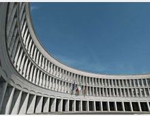 Facciata curva di un grande edificio istituzionale in cemento e vetro, con file regolari di finestre e pilastri verticali su più piani. Al centro sono visibili aste con bandiere davanti a un ampio spazio pavimentato sotto un cielo azzurro.