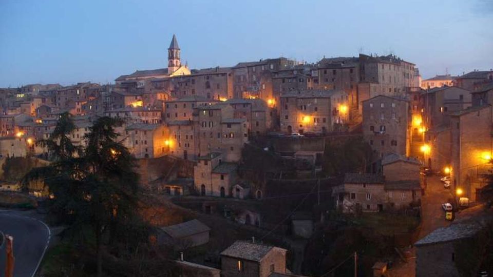 Panorama al crepuscolo di un borgo collinare con case in pietra addossate tra loro e un campanile che spicca sul profilo degli edifici. Luci stradali arancioni illuminano le facciate e le vie, con una strada curva e alberi in primo piano.