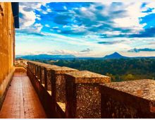 Terrazza panoramica di un edificio in pietra con lungo camminamento in mattoni e parapetto massiccio, affacciata su una vasta area boscosa. Sullo sfondo si vedono colline e una montagna isolata sotto un cielo ampio con nuvole.