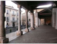 Portico storico con pavimentazione in pietra e lunga fila di colonne in muratura che sorreggono un soffitto con travi a vista. Sul lato aperto si affaccia su una strada e su edifici d’epoca con finestre e balconcini, separati da una ringhiera.