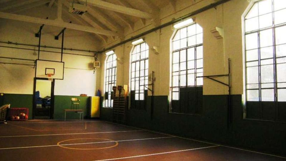 Palestra scolastica interna con campo da basket segnato a terra, soffitto alto con travi a vista e canestro fissato alla parete. Lungo un lato ci sono grandi finestre ad arco con inferriate che illuminano l’ambiente, con pareti bianche e fascia scura nella parte inferiore.