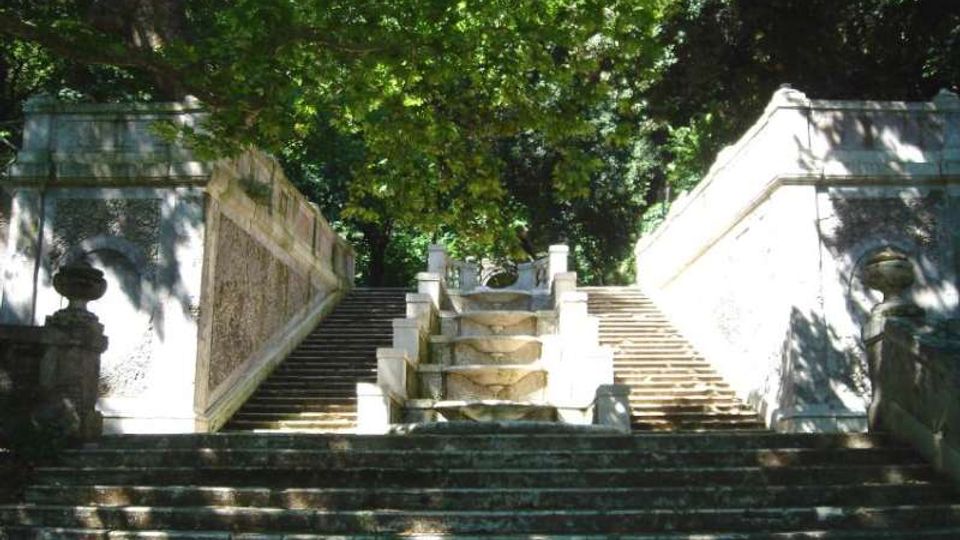 Scalinata in pietra all’aperto con rampe laterali simmetriche e un elemento centrale a gradini ondulati, fiancheggiata da parapetti in muratura con dettagli decorativi. L’area è circondata da alberi alti che creano ombra e luce filtrata sul percorso.