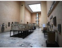 Sala museale con pareti alte color beige e lucernario rettangolare sul soffitto, pavimento in pietra e reperti lapidei esposti lungo le pareti. Al centro ci sono grandi bassorilievi su supporti metallici in fila e, in fondo, una colonna monumentale su piedistallo.