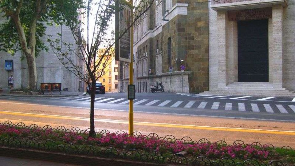 Vista di una strada urbana con attraversamento pedonale, un’aiuola con fiori rosa e piccoli alberi in primo piano. Sullo sfondo si vedono edifici in pietra e mattoni con grandi ingressi e finestre, lungo un incrocio.