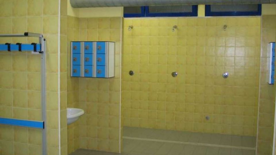 Spogliatoio con area docce, pareti rivestite di piastrelle gialle e pavimento grigio. Sono visibili un condotto di aerazione a soffitto, finestre alte con telaio blu, armadietti blu a parete e una panca metallica con appendiabiti.