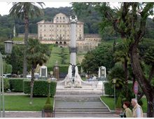 Vista di un viale monumentale in un giardino urbano con palme e siepi potate, che conduce a una grande colonna con sculture e scalinate. Sullo sfondo si vede un ampio edificio storico su una collina boscosa.