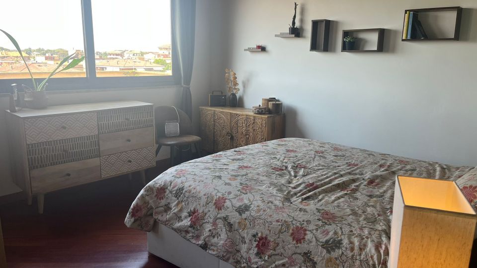 Camera da letto luminosa con grande finestra e tende grigie affacciata sui tetti della città, pavimento in legno scuro e letto con copriletto floreale. Sulla parete bianca ci sono mensole e cubi a muro, accanto a comò e credenza in legno decorato con piccoli oggetti e una lampada da tavolo.
