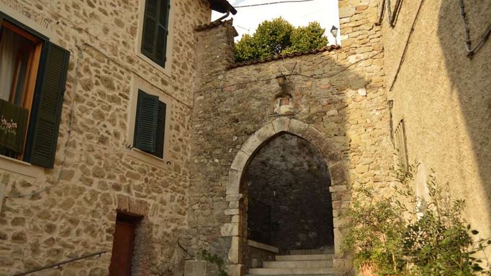 Vicolo in un borgo storico con edifici in pietra e persiane scure, che conduce a una scalinata. In fondo si vede un arco ogivale in muratura, simile a un’antica porta, incassato tra le pareti di pietra.