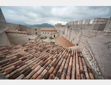 Veduta dall’alto dell’interno di una fortezza in pietra, con tetti in coppi di terracotta e mura merlate lungo il perimetro. Sullo sfondo si vedono montagne e cielo nuvoloso.