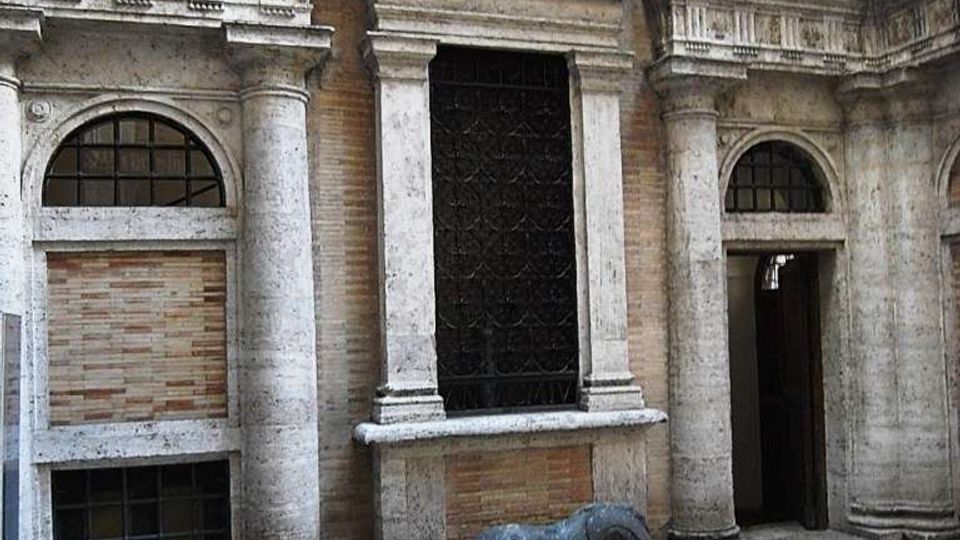 Cortile interno di un edificio storico con facciata in pietra e mattoni, colonne e archi, e una grande finestra con grata in ferro. Sul pavimento in pietra decorato è visibile una scultura sdraiata su un basamento, vicino a un ingresso laterale.