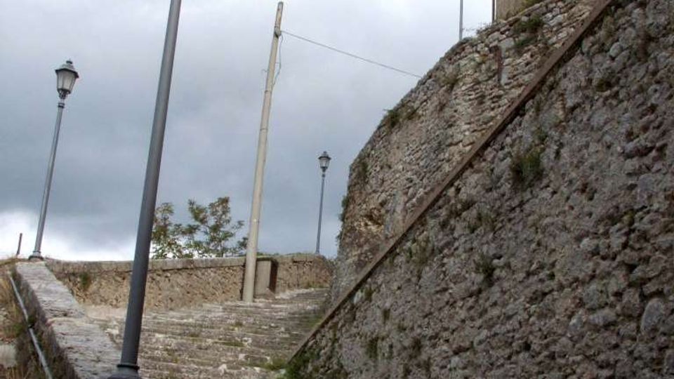 Scalinata in pietra che sale tra alti muri di contenimento in muratura grezza, affiancata da lampioni lungo il percorso. In cima si intravede un edificio con facciata intonacata e tetto spiovente sotto un cielo nuvoloso.