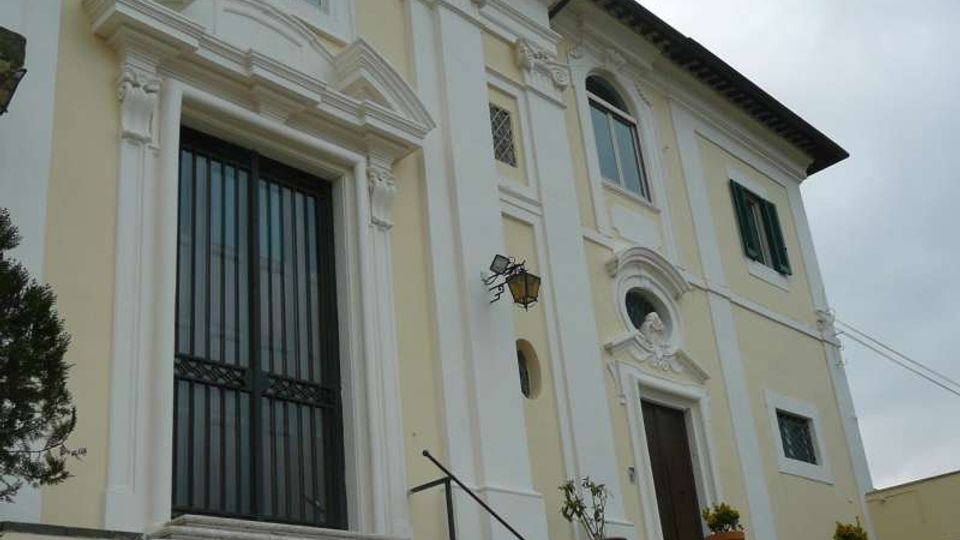 Facciata di un edificio storico in stile barocco color crema e bianco, con cornici decorative attorno a finestre e portali. Un’ampia scalinata in pietra conduce a un grande ingresso con cancello scuro, con piccoli vasi di piante ai lati e un campanile visibile sul tetto.