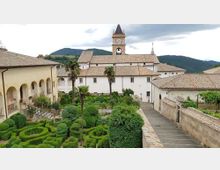 Cortile di complesso monastico con giardino all’italiana di siepi potate a labirinto e alcune palme, circondato da edifici chiari con tetti in coppi. Sullo sfondo si vede una torre dell’orologio e colline verdi sotto un cielo nuvoloso.