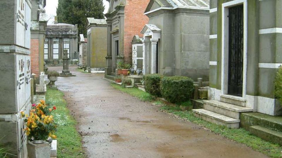 Viale di un cimitero con strada sterrata tra tombe e cappelle funerarie in muratura e pietra, alcune con colonne e porte scure. Ai lati si vedono piccoli cespugli, fiori in vaso e alti alberi sullo sfondo.