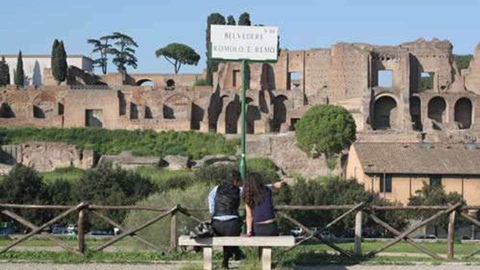 Panchina in un parco con staccionata in legno e cartello “ROMOLO E REMO” in primo piano, con vista su rovine archeologiche in muratura e archi sullo sfondo. Attorno si vedono alberi alti e vegetazione, con un tratto di strada e edifici bassi ai piedi delle rovine.