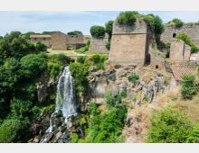 Cascata che scende da una scogliera rocciosa circondata da vegetazione fitta, accanto a mura e bastioni in pietra di una fortificazione storica. Sullo sfondo si vedono edifici in muratura e alberi su un altopiano sopra la gola.