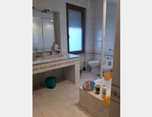 Bagno piastrellato in bianco con fascia decorativa, doppio lavabo con grande specchio e finestra con telaio in legno. A destra si vedono WC e bidet e una vasca con parete in vetro, con alcuni oggetti appoggiati sul bordo.