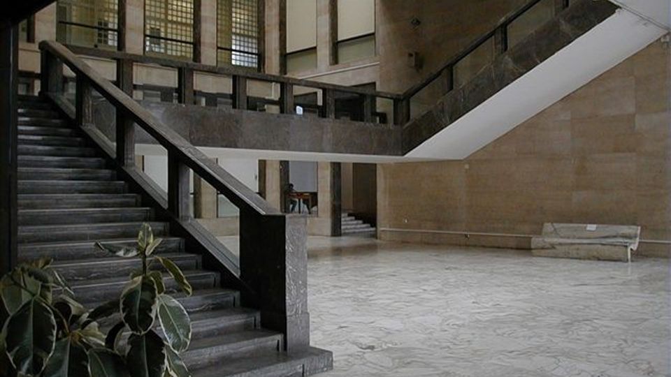 Atrio interno di un edificio con ampie scale in pietra e ringhiere scure, pavimento in marmo chiaro e pareti rivestite in lastre di pietra. Sullo sfondo si vedono grandi finestre a griglia e un ballatoio che corre lungo il piano superiore.