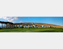 Panorama di un antico acquedotto romano in pietra con una lunga fila di arcate, esteso in orizzontale sopra un ampio prato verde. Sullo sfondo si vedono alberi ad alto fusto e un cielo azzurro con alcune nuvole.
