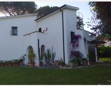 Casa in stile mediterraneo con pareti bianche e tetto spiovente, finestre con inferriate e una scala a chiocciola esterna che porta alla terrazza. Davanti c’è un prato con patio in pietra e vasi di cactus e altre piante, tra cui una bougainvillea viola che sale sulla facciata.