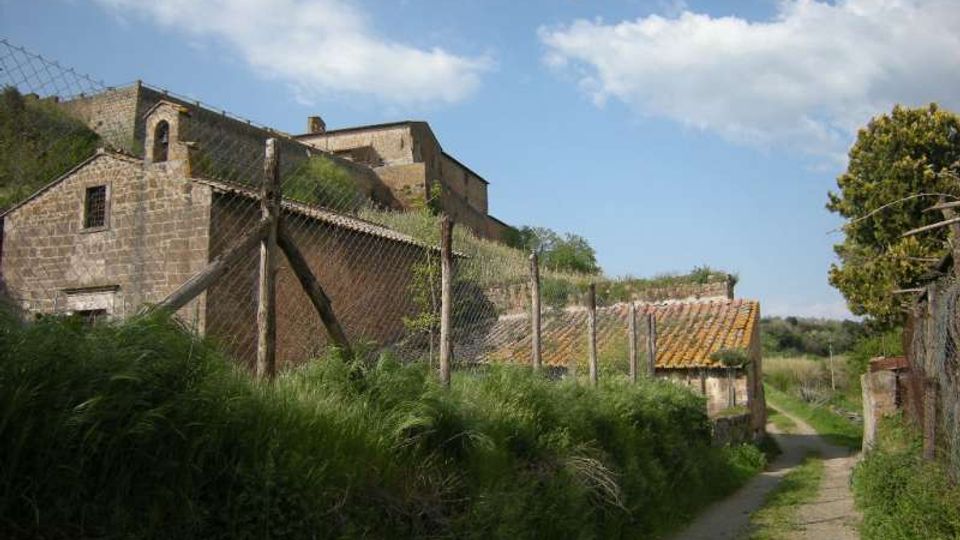 Strada di campagna stretta che costeggia una recinzione metallica, con un complesso di edifici in pietra e tetti in tegole su un pendio erboso. Sullo sfondo cielo azzurro con nuvole e vegetazione rurale ai lati del percorso.