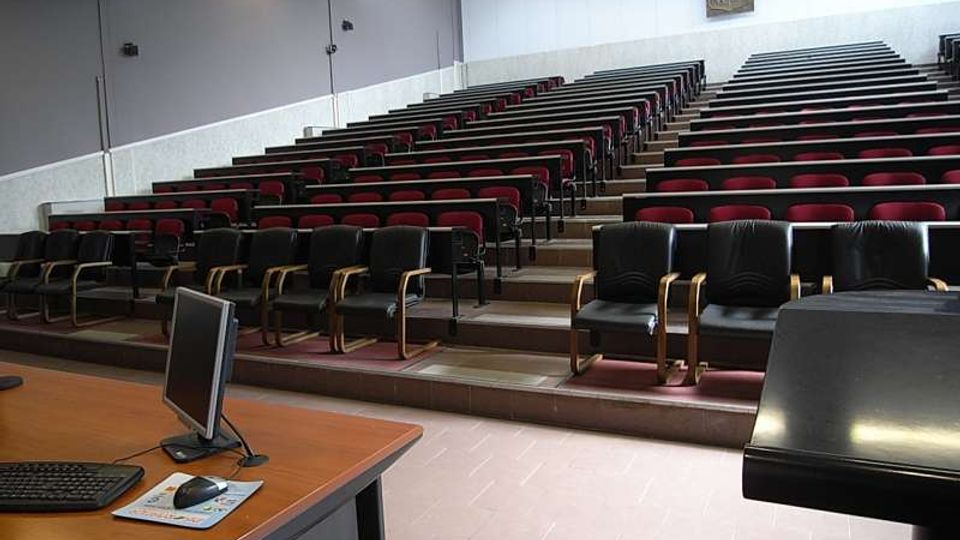 Aula universitaria con gradinate e file di sedili imbottiti rossi e neri disposti a livelli, con pareti chiare e illuminazione a soffitto. In primo piano si vede un banco docente con computer e, sul lato, un leggio nero.