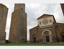 Facciata di una chiesa romanica in pietra con grande rosone centrale e portale ad arco, affiancata da un’alta torre medievale in muratura. Davanti si estende un prato, con cielo nuvoloso sullo sfondo.